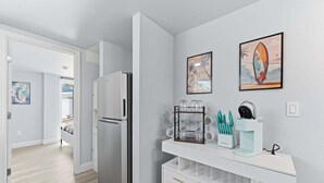 A modern kitchen with a refrigerator, a white countertop with glassware, and two framed artworks. - Walk To Beach, North End Restaurants, Night Life Steps Away! (Fort Myers Beach)