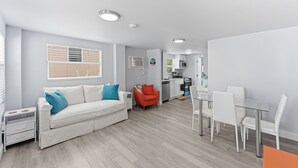 A modern living room with a sofa, a dining table with chairs, a kitchen area, and a small orange chair. - Walk To Beach, North End Restaurants, Night Life Steps Away! (Fort Myers Beach)