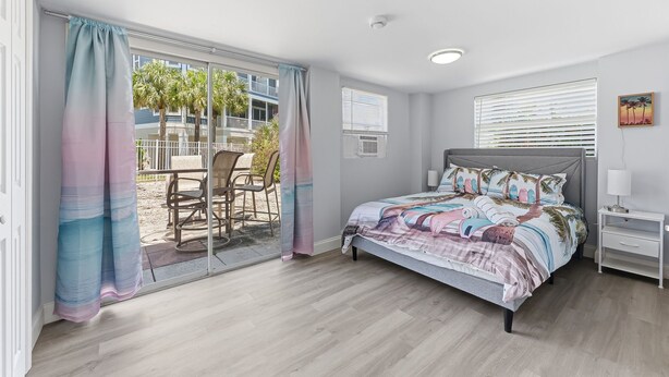 A bedroom with a bed, a nightstand, a window with blinds, and a view of palm trees outside. - Walk To Beach, North End Restaurants, Night Life Steps Away! (Fort Myers Beach)