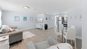 A modern living room with a sofa, a glass dining table, and a kitchen area in the background. - Walk To Beach, North End Restaurants, Night Life Steps Away! (Fort Myers Beach)