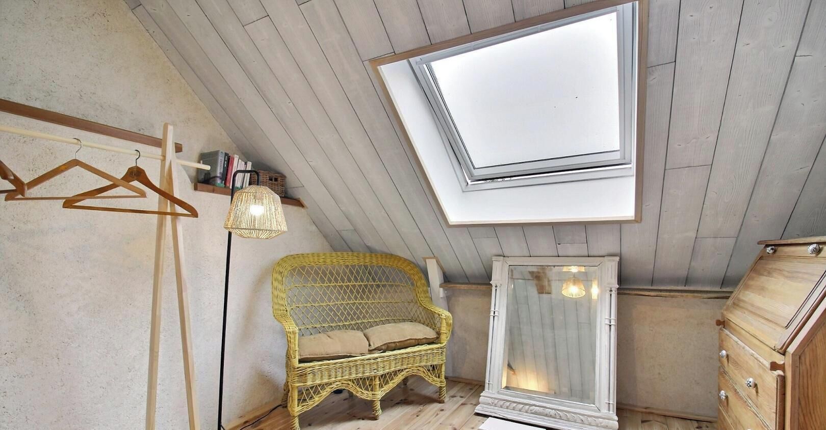 A room with a sloped ceiling, a wicker sofa, a dresser, a mirror, and a clothing rack.
