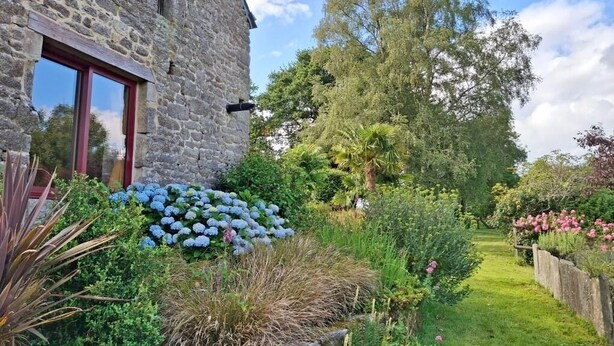 A stone building with a red-framed window, surrounded by lush greenery and flowering plants. - The owl shelter (LANGONNET)
