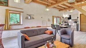 A spacious living area with a wooden ceiling, a sofa, chairs, a dining table, and a kitchen in the background. - The owl shelter (LANGONNET)
