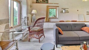A room with a wooden ceiling, a grey sofa, a wicker chair, a table with a lamp, and a painting on the wall. - The owl shelter (LANGONNET)