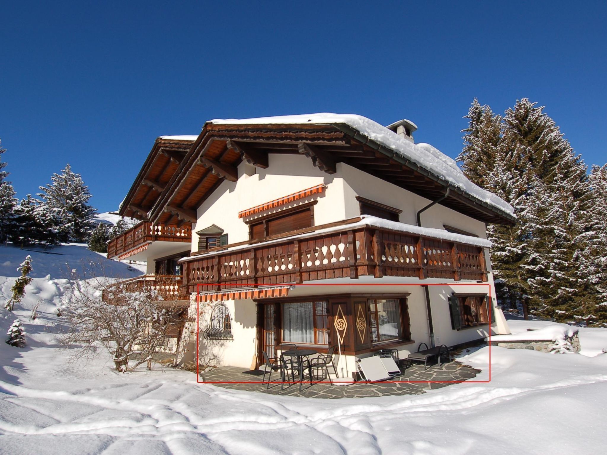 A chalet with a balcony, a stone patio, and snow-covered surroundings.