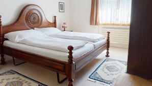 Een net opgemaakt bed in een kamer met een houten hoofdeinde, een klein nachtkastje met een lamp, een raam met gordijnen en een kamerbreed tapijt met patroon.