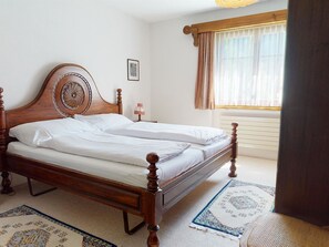 A neatly made bed in a room with a wooden headboard, a small bedside table with a lamp, a window with curtains, and a patterned rug on the floor. - Uehlinger Soland Appartement/Fewo, 0007 Soland Ferienwohnung Soland by Interhome (Lenzerheide)