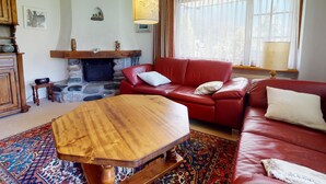 Een woonkamer met een rode leren bank, een houten salontafel, een stenen open haard en een groot raam met dunne gordijnen.