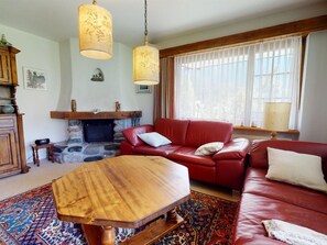 A living room with a red leather sofa, a wooden coffee table, a stone fireplace, and a large window with sheer curtains. - Uehlinger Soland Appartement/Fewo, 0007 Soland Ferienwohnung Soland by Interhome (Lenzerheide)