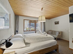 A bedroom with a wooden ceiling, a bed with white bedding, a window with a view of a snowy landscape, and a sofa with plaid cushions. - Riedblick Appartement/Fewo,   Ferienwohnung Riedblick by Interhome (Churwalden)