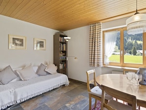 Um quarto aconchegante com teto de madeira, um sofá com almofadas, uma estante de livros, uma mesa de jantar com cadeiras e uma janela com vista para o exterior.