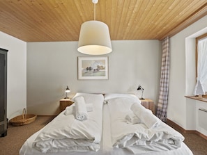 Uma cama bem arrumada com lençóis brancos, duas luminárias de cabeceira, um quadro na parede e teto de madeira.