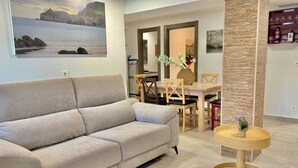 Una sala de estar con un sofá, una mesa de comedor con sillas, una mesita redonda de madera y un cuadro colgado en la pared.