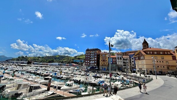 Un puerto deportivo con numerosas embarcaciones amarradas, un paseo peatonal y edificios de arquitectura variada.