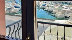 Vista a través de una ventana con barandilla de metal, cielo despejado y un puerto deportivo con barcos.