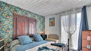 A bedroom with a wooden wardrobe, a bed with patterned bedding, a nightstand, and a window with curtains. - Géraldine - LE PALAIS - Gîte (LE PALAIS)