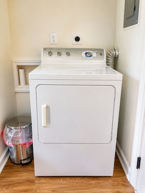 A white front-loading washer and dryer in a laundry room with a wooden floor, a metal trash can, and a white shelf.