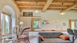 A room with a wooden ceiling, a grey sofa, a wicker chair, a table with a lamp, and a painting on the wall. - The bird sanctuary (LANGONNET)