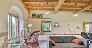 Ein Zimmer mit Holzboden, einem grauen Sofa, einem Korbsessel, einem Tisch mit Lampe und einem Bild an der Wand.