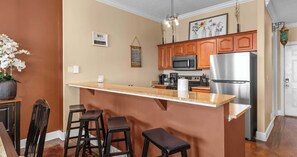 A kitchen with wooden cabinets, a stainless steel refrigerator, a microwave, and a bar area with stools. - Above the Waves Oceanfront (Panama City Beach)