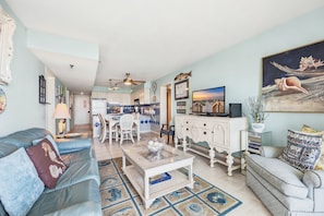 Ein Wohnzimmer mit Sofa, Couchtisch und Fernseher. Im Hintergrund ist eine Küche mit weißen Schränken und blauen Fliesen zu sehen.