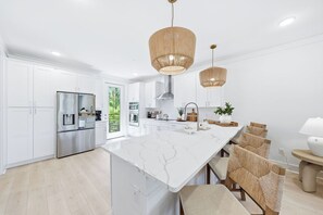 A modern kitchen with white cabinetry, a large island, stainless steel appliances, and a dining area with wicker chairs. - Luxury, Blue Eden Bliss Sleeps 12, Close to Bay (Santa Rosa Beach)