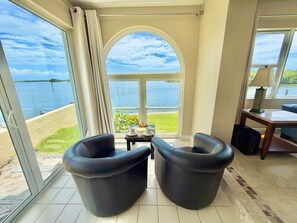 A room with a large window overlooking a beach and water, two black chairs, a small table with a cup and saucer, and a lamp on a side table. - Spanish Trace Escape | Pool + Minutes to the Beach (Cocoa Beach)