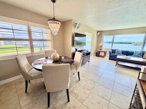 A spacious dining area with a round table and chairs, a chandelier, and a view of the water through large windows. - Spanish Trace Escape | Pool + Minutes to the Beach (Cocoa Beach)