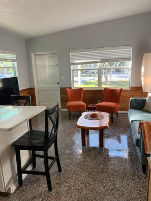 Un espacio habitable compacto con kitchenette, zona de comedor con dos sillas, sofá y ventana con persianas.