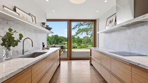 A modern kitchen with marble countertops, wooden cabinets, and a view of a deck and greenery. - River View by Avantstay Arch Digest 1863 Italianate + Pool + Screening Room Hudson Views (Germantown)