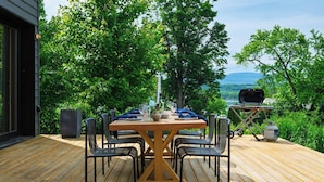 An outdoor dining area with a wooden table and chairs, surrounded by lush greenery and a view of a lake. - River View by Avantstay Arch Digest 1863 Italianate + Pool + Screening Room Hudson Views (Germantown)