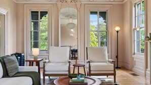 A spacious room with two white armchairs, a sofa, a small table with books, and a floor lamp. Large windows provide natural light and a view of greenery outside. - River View by Avantstay Arch Digest 1863 Italianate + Pool + Screening Room Hudson Views (Germantown)