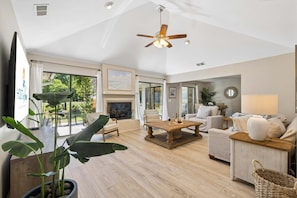 A spacious living room with a fireplace, comfortable seating, a ceiling fan, and large windows offering a view of the outdoors. - Sea Pines 3BR, Pet-Friendly, South Beach Pool Access, Bike to Beach & Golf! (Hilton Head Island)