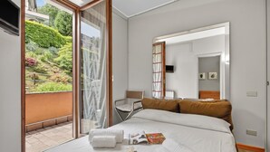Ett hotellrum med en säng, en stol, ett litet bord och en skjutdörr i glas som leder ut till en balkong med utsikt över en trädgård.