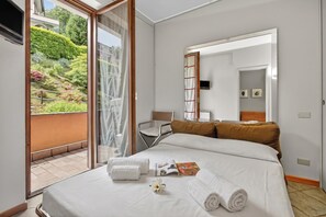 A hotel room with a bed, a chair, a small table, and a sliding glass door leading to a balcony with a garden view. - Riflessi Di Luce: Lake Maggiore View Balcony Stresa, Stresa, Italy (Stresa)