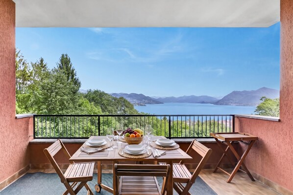 A balcony with a table set for two, a bowl of fruit, and a view of a lake and mountains. - Riflessi Di Luce: Lake Maggiore View Balcony Stresa, Stresa, Italy (Stresa)