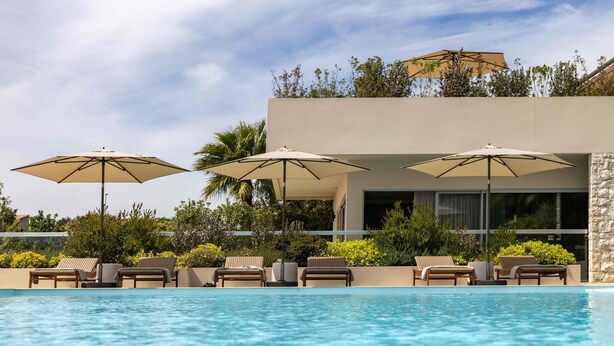 Een zwembadgedeelte met ligbedden en parasols, een modern gebouw met een stenen gevel, en een heldere lucht.
