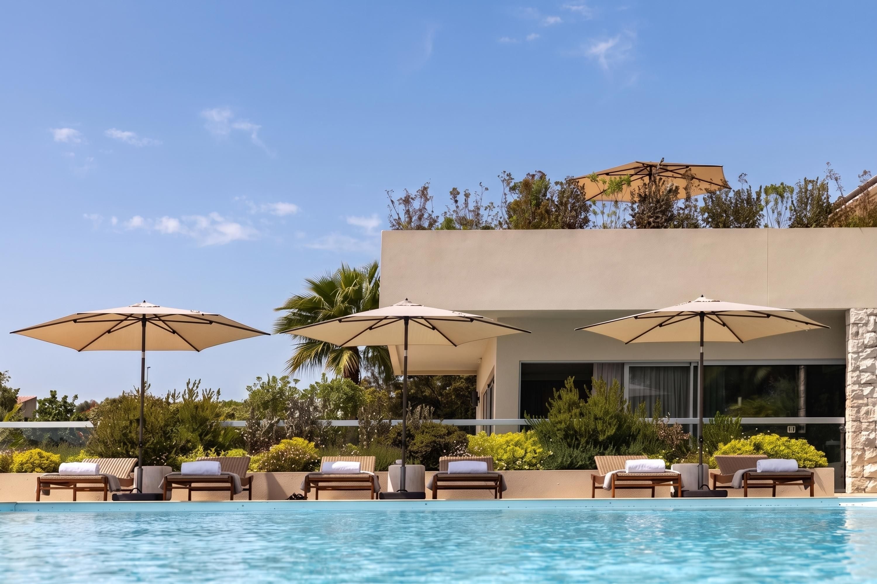 A poolside area with lounge chairs and umbrellas, a clear blue sky, and a modern building with a stone facade.