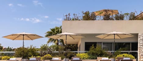A poolside area with lounge chairs and umbrellas, a clear blue sky, and a modern building with a stone facade.