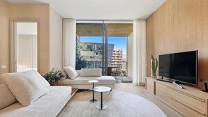 Un salon moderne avec un canapé, deux petites tables, un téléviseur et une grande fenêtre donnant sur des immeubles.