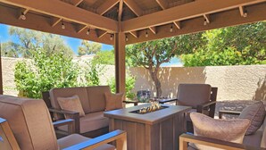 Patio esterno con tetto in legno, un tavolo centrale dotato di focolare e diverse zone salotto con cuscini.