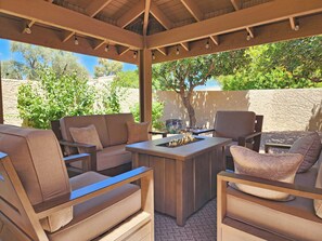 Eine Terrasse im Freien mit Holzboden, einem zentralen Tisch mit Feuerstelle und mehreren Sitzbereichen mit Kissen.