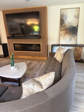 Ein modernes Wohnzimmer mit einem großen Flachbildfernseher, einem grauen Sofa mit gemusterten Kissen, einem kleinen weißen Tisch mit einem Kaktus und einem großen abstrakten Gemälde an der Wand.