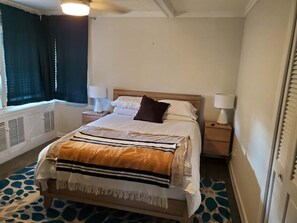 Ein Hotelzimmer mit einem Bett, zwei Nachttischlampen, einem Fenster mit Vorhängen, einer Heizung und einem gemusterten Teppich.