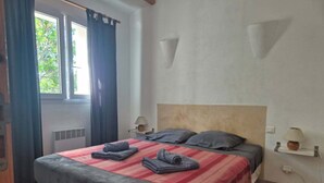 A bedroom with a bed, two bedside lamps, a window with curtains, and a wall-mounted shelf.