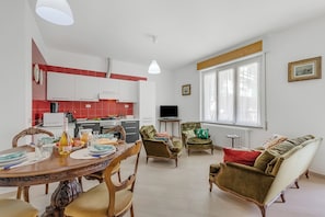 Eine moderne Küche mit weißen Schränken, einem Esstisch gedeckt mit Tellern, Gläsern und einer Flasche, sowie ein Wohnbereich mit Sofa und Sesseln.