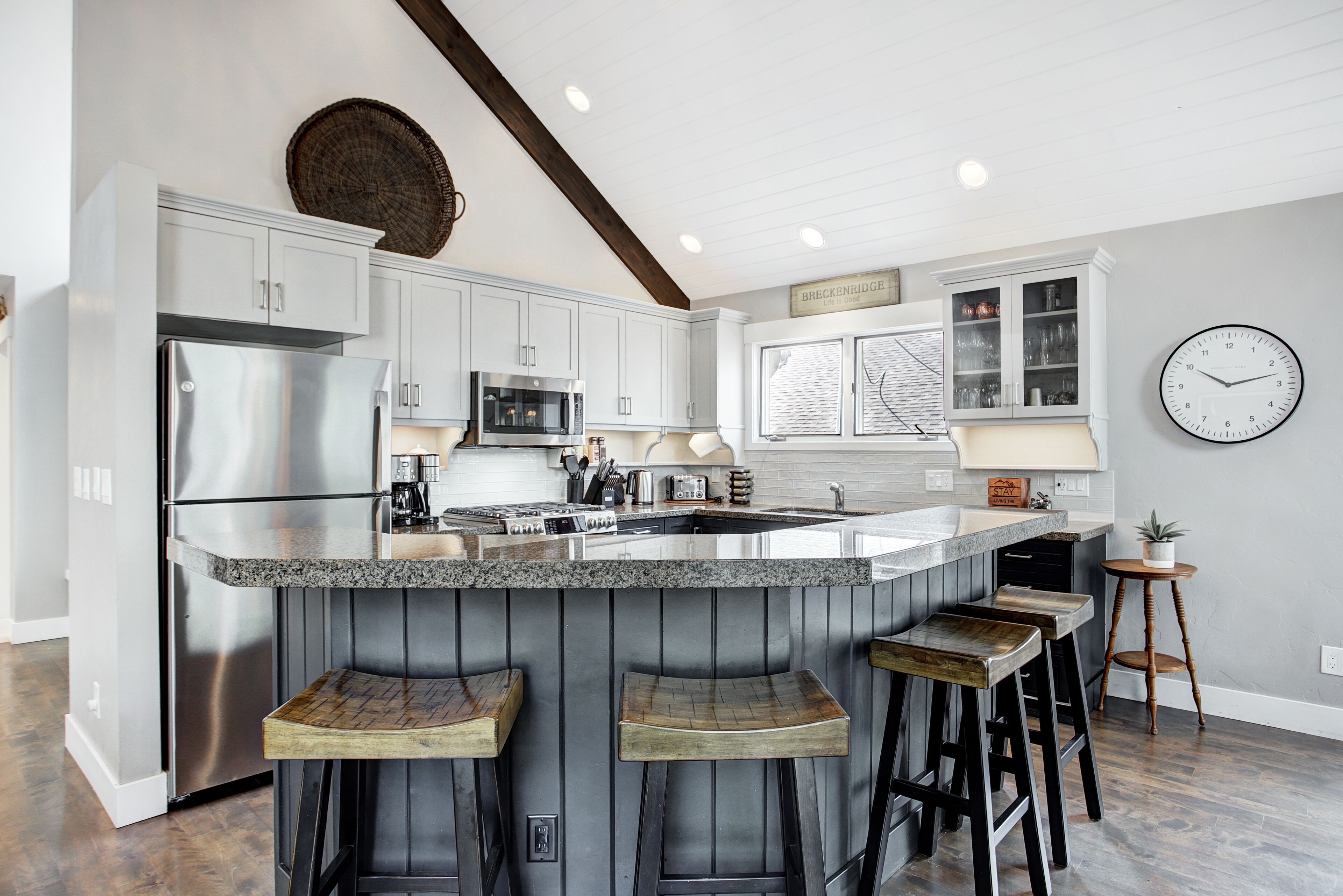 Una cocina con una isla central, taburetes de bar, refrigerador, microondas y un reloj en la pared.