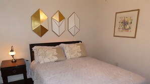 Ein ordentlich bezogenes Bett mit blumigen Kissen, ein Nachttisch mit Lampe und drei geometrische Wandbilder.