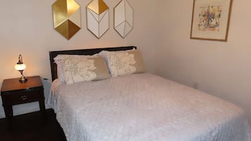 Un lit bien fait, agrémenté de coussins à motifs floraux, une table de chevet avec une lampe, et trois œuvres d’art géométriques accrochées au mur.