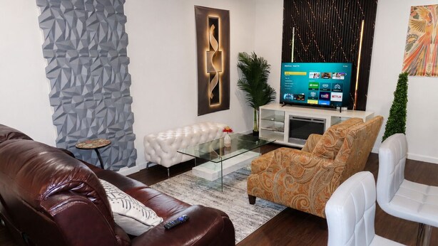 Ein modernes Wohnzimmer mit Ledersofa, gemustertem Sessel und Glastisch. Ein Flachbildfernseher ist an der Wand montiert, und links befindet sich eine dekorative Wandverkleidung.